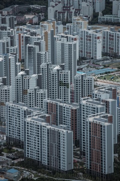 A densely packed urban landscape with numerous high-rise residential buildings. The structures are predominantly rectangular, featuring a mix of gray, white, and subtle earth tones. The perspective is from above, showcasing the organized grid layout typical of a cityscape. Parks and small roads can be seen between the buildings.