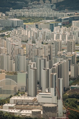 A dense urban landscape filled with numerous high-rise residential buildings and skyscrapers. The structures exhibit modern architectural designs, predominantly in white and gray tones. In the foreground, there is a construction crane and several industrial facilities with large cylindrical tanks. The background features more buildings extending up to a hilly, green horizon.