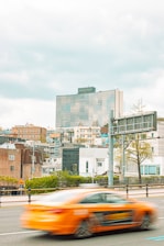 A cityscape featuring a modern high-rise building with the 'Grand Hyatt' name on top, surrounded by various residential and commercial structures. A bright orange car, slightly blurred, is captured in motion on the road in the foreground with a large traffic sign and some trees visible.