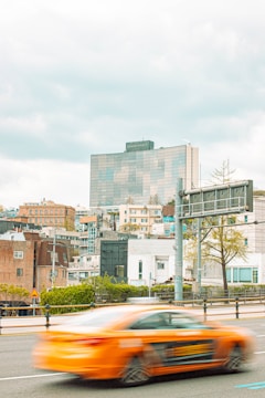A cityscape featuring a modern high-rise building with the 'Grand Hyatt' name on top, surrounded by various residential and commercial structures. A bright orange car, slightly blurred, is captured in motion on the road in the foreground with a large traffic sign and some trees visible.