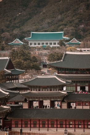 A complex of traditional Asian architectural buildings with green-tiled roofs and intricate wooden designs is seen, set against a backdrop of forested hills. There are several large structures, some with multiple tiers, and numerous people can be observed around and inside the complex.