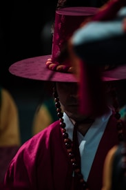 A person wearing traditional Korean attire including a distinctive tall hat and beads. The attire is vibrant, with a deep red hue that stands out against the darker background. The facial expression is partially obscured by shadows, adding a sense of mystery.