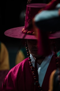 A person wearing traditional Korean attire including a distinctive tall hat and beads. The attire is vibrant, with a deep red hue that stands out against the darker background. The facial expression is partially obscured by shadows, adding a sense of mystery.
