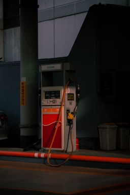 A gas pump stands in a dimly lit area with a yellow hose draped over it. The surroundings include a concrete pillar, a nearby parked car, and two trash bins. The area appears empty and quiet.