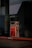 A gas pump stands in a dimly lit area with a yellow hose draped over it. The surroundings include a concrete pillar, a nearby parked car, and two trash bins. The area appears empty and quiet.