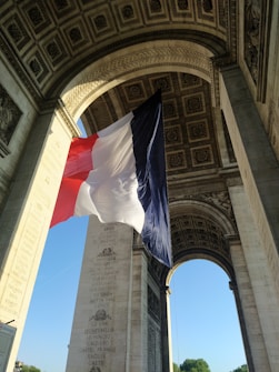 A large French flag is suspended under the arch of a monumental structure with intricate carvings and inscriptions. The arch's interior showcases detailed patterns and ornate designs.
