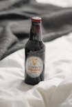 A bottle of Guinness Extra Stout is placed on a soft white fabric with a dark material in the background. The bottle has a distinct label featuring the brand's logo and is capped with a red top.
