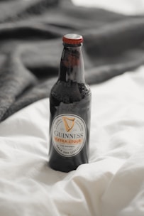 A bottle of Guinness Extra Stout is placed on a soft white fabric with a dark material in the background. The bottle has a distinct label featuring the brand's logo and is capped with a red top.