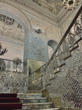 An ornate and luxurious interior featuring intricately detailed walls and ceilings with elaborate patterns and designs. A grand staircase with reflective surfaces and embellished balustrades leads upwards, while a chandelier adds to the opulence. The overall architecture includes decorative arches and extensive use of mirrors or glass elements that enhance the grandeur of the space.