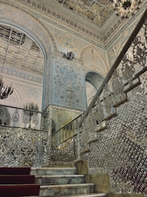 An ornate and luxurious interior featuring intricately detailed walls and ceilings with elaborate patterns and designs. A grand staircase with reflective surfaces and embellished balustrades leads upwards, while a chandelier adds to the opulence. The overall architecture includes decorative arches and extensive use of mirrors or glass elements that enhance the grandeur of the space.