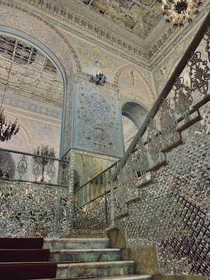 An ornate and luxurious interior featuring intricately detailed walls and ceilings with elaborate patterns and designs. A grand staircase with reflective surfaces and embellished balustrades leads upwards, while a chandelier adds to the opulence. The overall architecture includes decorative arches and extensive use of mirrors or glass elements that enhance the grandeur of the space.
