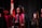 A group of graduates in black caps and gowns, with maroon and gold stoles. They appear to be participating in a ceremony, possibly on stage with a national flag and red floral decorations in the background.