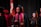 A group of graduates in black caps and gowns, with maroon and gold stoles. They appear to be participating in a ceremony, possibly on stage with a national flag and red floral decorations in the background.