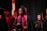 A group of graduates in black caps and gowns, with maroon and gold stoles. They appear to be participating in a ceremony, possibly on stage with a national flag and red floral decorations in the background.