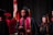 A group of graduates in black caps and gowns, with maroon and gold stoles. They appear to be participating in a ceremony, possibly on stage with a national flag and red floral decorations in the background.