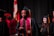 A group of graduates in black caps and gowns, with maroon and gold stoles. They appear to be participating in a ceremony, possibly on stage with a national flag and red floral decorations in the background.
