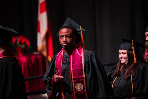A group of graduates in black caps and gowns, with maroon and gold stoles. They appear to be participating in a ceremony, possibly on stage with a national flag and red floral decorations in the background.