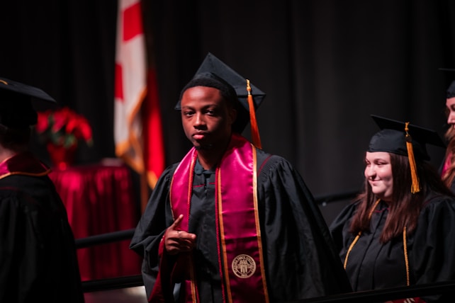 A group of graduates in black caps and gowns, with maroon and gold stoles. They appear to be participating in a ceremony, possibly on stage with a national flag and red floral decorations in the background.