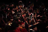 A large group of graduates wearing black caps and gowns are seated in an auditorium. Some are smiling and looking at their phones, while others are engaged in conversation. The atmosphere seems lively and filled with anticipation.