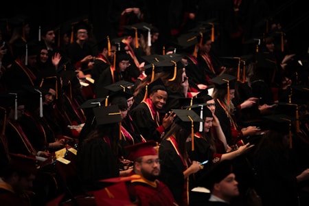A large group of graduates wearing black caps and gowns are seated in an auditorium. Some are smiling and looking at their phones, while others are engaged in conversation. The atmosphere seems lively and filled with anticipation.