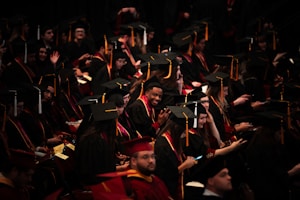 A large group of graduates wearing black caps and gowns are seated in an auditorium. Some are smiling and looking at their phones, while others are engaged in conversation. The atmosphere seems lively and filled with anticipation.