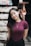 A young woman with long dark hair stands indoors, wearing a maroon shirt and a necklace with a cross pendant. She is smiling softly against a blurred background featuring shelves and a hanging decoration.