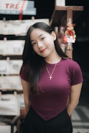 A young woman with long dark hair stands indoors, wearing a maroon shirt and a necklace with a cross pendant. She is smiling softly against a blurred background featuring shelves and a hanging decoration.