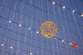 A decorative setup featuring numerous strings of small, twinkling lights hanging vertically against the evening sky. A large, illuminated spherical figure made of lights is prominently displayed, along with a red star-shaped decoration.