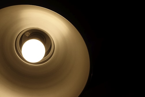 A close-up view of a lit lamp emitting a white glow, surrounded by a metallic, circular lampshade creating a gradient of light to dark as it extends outwards. The surrounding area is dark, emphasizing the brightness of the lamp.