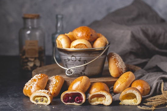A rustic setting featuring a metal bucket filled with various freshly baked bread rolls, displayed on a wooden surface. Several rolls are sliced open at the front to reveal different fillings, such as cherry, custard, and chocolate. Accompanying the scene are textured fabrics and glass jars, creating a warm, homely atmosphere.