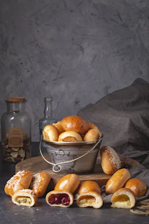 A collection of freshly baked bread rolls with various fillings is displayed. The rolls are arranged in a rustic metal bucket, with some spilling out onto a wooden surface. A few rolls are cut open to reveal different fillings, including a dark red berry filling, a creamy filling, and a seeded loaf. The setting includes a jar filled with preserved foods and a small bottle, providing a homely and traditional kitchen feel.