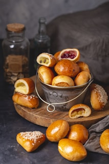 A rustic setting featuring a metal bowl filled with various pastries, some with fruit or chocolate fillings. The pastries are arranged on a wooden board, some topped with sesame seeds. There are glass jars and bottles in the background, giving an artisanal feel.