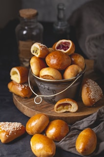 A rustic wooden board holds a metal bucket filled with a variety of small, golden-brown pastries. Some pastries are topped with sesame seeds or filled with different ingredients such as jam or custard. Scattered pastries surround the bucket, with a gray cloth draped nearby and glass bottles in the background.