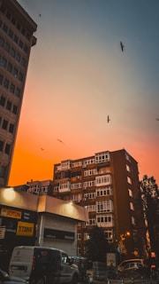 A cityscape at sunset with a striking orange and blue sky. Birds are flying above the tall buildings, which include an office and residential block. Streetlights are beginning to glow, and a few vehicles are parked on the street below. The storefronts display various signs, indicating a commercial area.