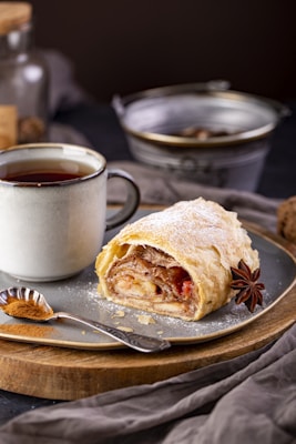 A slice of pastry, likely an apple strudel, dusted with powdered sugar on a plate. Next to it is a cup of tea or coffee in a ceramic mug. There is an anise star and a small spoon with cinnamon powder on the plate. In the background, there are blurred objects including a metal container and a glass jar, adding a cozy, homey atmosphere.