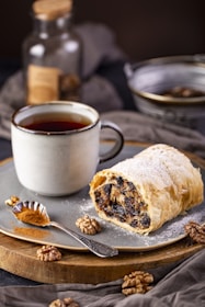 A rustic plate features a slice of pastry filled with dried fruits and nuts, dusted with powdered sugar. Beside it is a steaming cup of tea or coffee in a ceramic mug. A small spoon filled with ground cinnamon rests on the plate, with whole walnuts scattered around. The soft lighting and warm tones suggest a cozy, inviting setting.