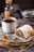 A rustic plate features a slice of pastry filled with dried fruits and nuts, dusted with powdered sugar. Beside it is a steaming cup of tea or coffee in a ceramic mug. A small spoon filled with ground cinnamon rests on the plate, with whole walnuts scattered around. The soft lighting and warm tones suggest a cozy, inviting setting.