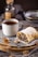 A rustic plate features a slice of pastry filled with dried fruits and nuts, dusted with powdered sugar. Beside it is a steaming cup of tea or coffee in a ceramic mug. A small spoon filled with ground cinnamon rests on the plate, with whole walnuts scattered around. The soft lighting and warm tones suggest a cozy, inviting setting.