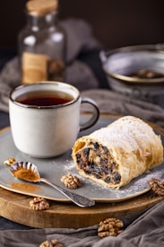 A rustic plate features a slice of pastry filled with dried fruits and nuts, dusted with powdered sugar. Beside it is a steaming cup of tea or coffee in a ceramic mug. A small spoon filled with ground cinnamon rests on the plate, with whole walnuts scattered around. The soft lighting and warm tones suggest a cozy, inviting setting.