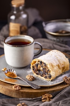 A rustic plate features a slice of pastry filled with dried fruits and nuts, dusted with powdered sugar. Beside it is a steaming cup of tea or coffee in a ceramic mug. A small spoon filled with ground cinnamon rests on the plate, with whole walnuts scattered around. The soft lighting and warm tones suggest a cozy, inviting setting.