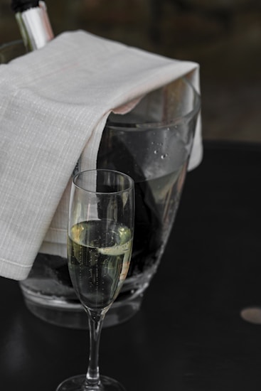 A glass filled with champagne or sparkling wine is placed in the foreground on a dark surface. Behind it, a large ice bucket holds a bottle wrapped with a white cloth, suggesting a chilled beverage. The background is slightly out of focus, possibly depicting a subtle dark setting, enhancing the elegance of the scene.