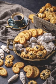An assortment of pastries and cookies is artfully arranged on a dark textured surface. A mix of powdered sugar-coated confections, buttery cookies, and intricate designs are present. A cup of black coffee sits in a greenish cup and saucer, adding a contrasting element to the image. The scene evokes an inviting and cozy atmosphere.
