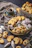 An assortment of pastries and cookies is artfully arranged on a dark textured surface. A mix of powdered sugar-coated confections, buttery cookies, and intricate designs are present. A cup of black coffee sits in a greenish cup and saucer, adding a contrasting element to the image. The scene evokes an inviting and cozy atmosphere.