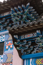 Intricate and colorful architectural details with traditional Asian motifs, featuring vibrant patterns and curved, interlocking wooden elements. The design includes geometric shapes and ornamental decorations with vivid blue, green, and orange hues.