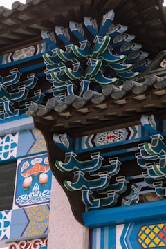 Intricate and colorful architectural details with traditional Asian motifs, featuring vibrant patterns and curved, interlocking wooden elements. The design includes geometric shapes and ornamental decorations with vivid blue, green, and orange hues.