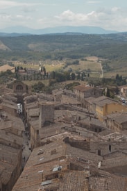 A view of a historic town with old, stone buildings and clay-tiled roofs. Narrow streets wind through the town, surrounded by a landscape of vineyards and rolling hills. In the distance, the lush greenery of trees and fields stretches towards a mountainous horizon under a partly cloudy sky.