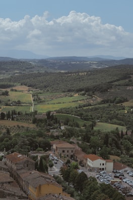 A picturesque landscape featuring a small town with rustic buildings, surrounded by lush greenery and vineyards. Rolling hills stretch into the distance under a vast sky with fluffy clouds.