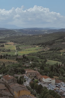A picturesque landscape featuring a small town with rustic buildings, surrounded by lush greenery and vineyards. Rolling hills stretch into the distance under a vast sky with fluffy clouds.