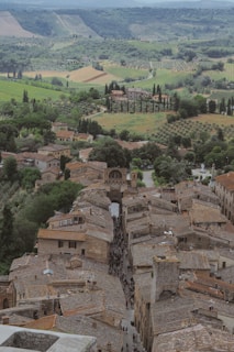 A charming medieval village with narrow cobblestone streets and rustic terracotta-roofed buildings. The village is set amidst rolling green hills, vineyards, and tall cypress trees, offering a picturesque view of the countryside.