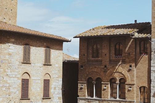 Two rustic buildings feature classic Italian architecture, with arched windows and terracotta roof tiles. The stone facades display a mix of earthy tones under a clear blue sky.